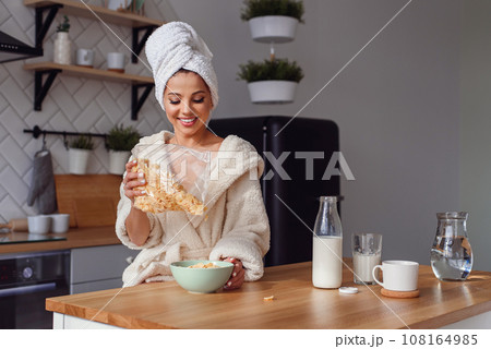 Charming woman in a bathrobe and towel on her head prepares a healthy breakfast with flakes and milk. The girl has a breakfast on stylish cozy kitchen at the morning. 108164985