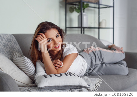 Pretty young woman with beautiful smile lies on grey couch with pillows at cozy home. Cute girl having rest at home in lazy day. 108165220