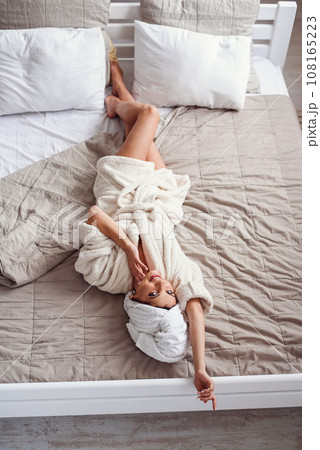 A pretty happy young woman lying on the bed at home after bath dressed in white bathrobe. Beautiful girl having relax on the bed in morning. Loking on the camera. 108165223
