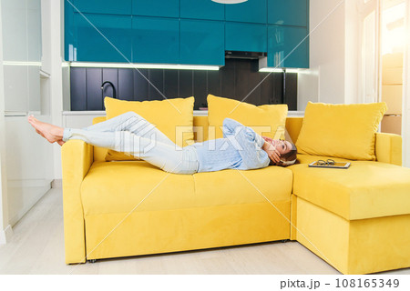 Pretty blonde teenage girl in headphones is listening to music, looking at camera and smiling while lying on her back, legs up, on sofa at home Pretty blonde teenage girl in headphones is listening to music, looking at camera and smiling while lying on her back, legs up, on sofa at home 108165349