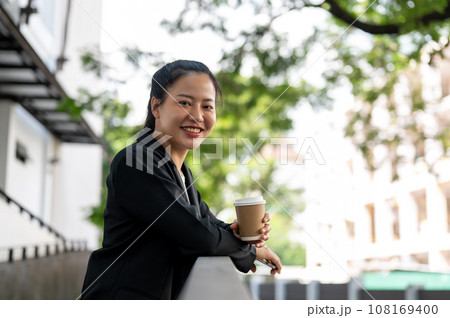 A cheerful and beautiful Asian businesswoman is relaxing outdoors during her coffee break. A cheerful and beautiful Asian businesswoman is relaxing outdoors during her coffee break. 108169400