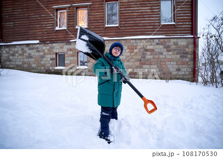 A boy in a green down jacket with a shovel stands in the yard in winter. 108170530