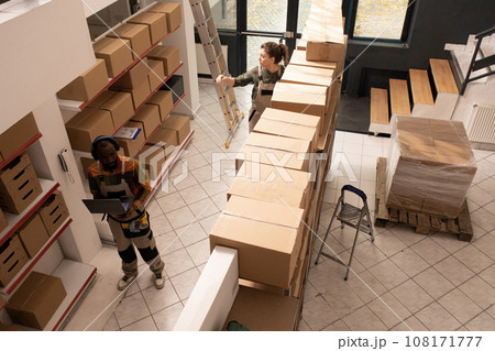 Top view of diverse coworkers working in warehouse, preparing customers orders before start shipping merchandise. Storage room employees in protective overall working with cardboard boxes Top view of diverse coworkers working in warehouse, preparing customers orders before start shipping merchandise. Storage room employees in protective overall working with cardboard boxes 108171777