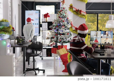 Businessman in teleconference meeting with coworkers sitting at desk in Christmas decorated workplace. Merry employee wearing festive seasonal clothing chatting with colleagues in online videocall Businessman in teleconference meeting with coworkers sitting at desk in Christmas decorated workplace. Merry employee wearing festive seasonal clothing chatting with colleagues in online videocall 108171811