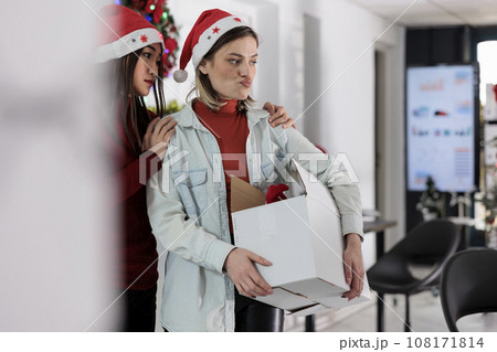 Asian employee upset by company manager leaving Christmas decorated office after deciding to quit. Team leader holding desk belongings box, bidding farewell to assistant before starting another job 108171814