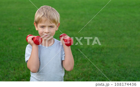 Sporty little boy lifting dumbbells on a green lawn. Joyful toddler doing exercise against nature background outdoors. Happy Child holding red dumbbells, looking at camera. Sport for kids concept. Sporty little boy lifting dumbbells on a green lawn. Joyful toddler doing exercise against nature background outdoors. Happy Child holding red dumbbells, looking at camera. Sport for kids concept. 108174436