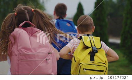 Active schoolchildren run carrying schoolbags on city street. Happy little friends with backpacks run together to school across park. Little children hurry to school running along city street Active schoolchildren run carrying schoolbags on city street. Happy little friends with backpacks run together to school across park. Little children hurry to school running along city street 108177324