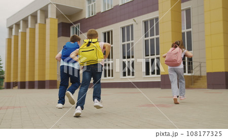 Playful children with schoolbags run together to school building backside view. Little students with backpacks hurry on school lessons. Active little children run to school in morning Playful children with schoolbags run together to school building backside view. Little students with backpacks hurry on school lessons. Active little children run to school in morning 108177325