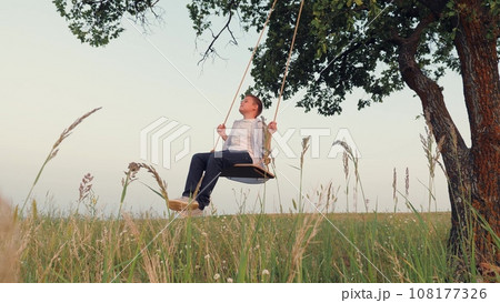 Dreaming preteen schoolboy plays swings under high tree at sunset light. Smiling little boy swings resting in country field on summer holiday. Little boy rides handmade swing in evening country garden Dreaming preteen schoolboy plays swings under high tree at sunset light. Smiling little boy swings resting in country field on summer holiday. Little boy rides handmade swing in evening country garden 108177326