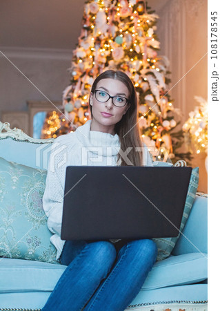 Cute lady in white sweater using laptop sitting on blue sofa in front of the Christmas tree at home 108178545