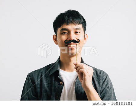 A joyful Asian man in a portrait holds a funny mustache card, radiating happiness and humor. Isolated on white, it's a Fathers Day and November-themed image. 108181181