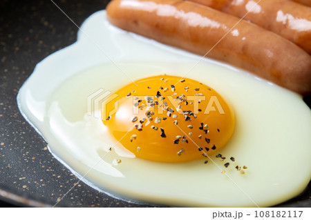 目玉焼き(粗びきブラックペッパーがけ)とウインナーソーセージをフライパンで焼く調理シーン。 目玉焼き(粗びきブラックペッパーがけ)とウインナーソーセージをフライパンで焼く調理シーン。 108182117