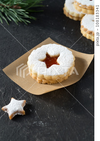 Yummy homemade Linzer cookies with jam and sugar powder on paper for baking. Dark textured background with pine branch. 108183341