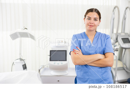 Young woman cosmetologist standing with crossed hands, smiling and looking at camera in clinic of aesthetic medicine Young woman cosmetologist standing with crossed hands, smiling and looking at camera in clinic of aesthetic medicine 108186517