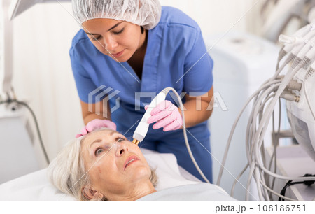 Elderly woman at appointment with beautician doing ultrasonic face cleaning with help of high-frequency ultrasonic waves in cosmetology clinic 108186751