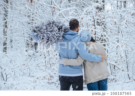 a couple hugs in a snowy forest. the man has a Christmas tree on his shoulder. back view 108187565