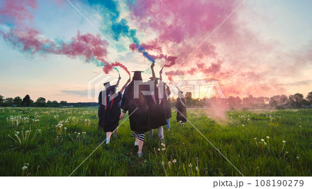 Graduates in costume walk across a field with coloured red and blue smoke at sunset. 108190279