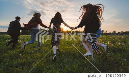 Cheerful friends run to meet the sun holding hands. 108190349