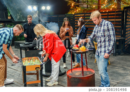 Close up of joyful guy with blond girl plays table football game while their male friend grilling vegetables in outdoors at weekend. Slow motion 108191498