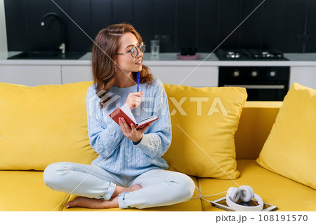 Attractive female writer in glasses writes her personal records into notebook while sitting in comfortable yellow couch in stylish modern kitchen. 108191570
