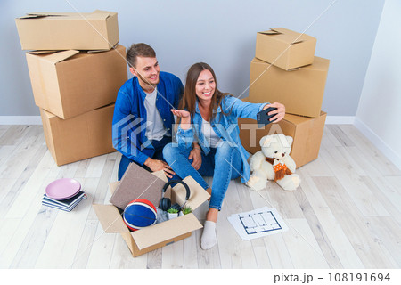 Young couple taking selfies in their new home while sitting among cardboard boxes. Happy attractive young couple is moving, making a selfie, cuddling and smiling. Young couple taking selfies in their new home while sitting among cardboard boxes. Happy attractive young couple is moving, making a selfie, cuddling and smiling. 108191694