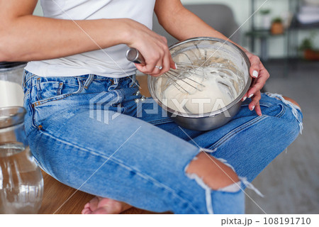 Hands of young woman in casual clothes kneads a dough in a metal plate at stylish kitchen. Young pretty woman baking on bright home kitchen. 108191710