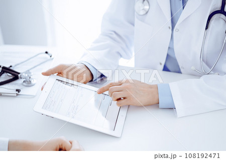 Doctor and patient sitting opposite each other at the desk in clinic. The focus is on female physician's hands pointing into tablet computer touchpad, close up. Medicine concept Doctor and patient sitting opposite each other at the desk in clinic. The focus is on female physician's hands pointing into tablet computer touchpad, close up. Medicine concept 108192471