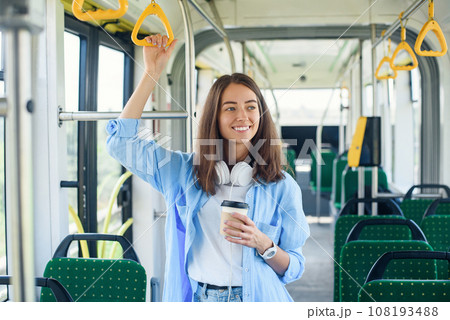Stylish woman in blue shirt enjoying trip in the modern tram or bus, stands with cup of coffee in the public transport. 108193488