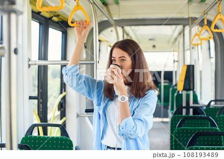 Beautiful woman in blue shit drinks coffee while riding in the public transport. Beautiful woman in blue shit drinks coffee while riding in the public transport. 108193489