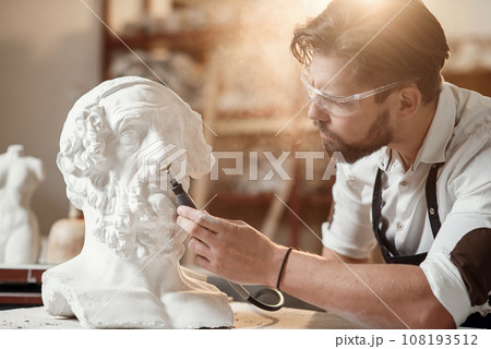 Professional bearded carver polishing with micro drill machine details of the man's sculpture head in his workshop. 108193512