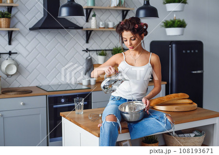 Beautiful smiling woman pours water in a deep metal plate to kneads a dough while sitting on the table in the modern stylish kitchen. 108193671