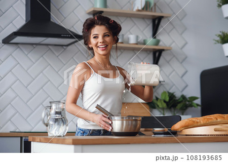 Cute young woman pours flour in a deep metal plate to kneads a dough while sitting on the table in the modern stylish kitchen. Cute young woman pours flour in a deep metal plate to kneads a dough while sitting on the table in the modern stylish kitchen. 108193685