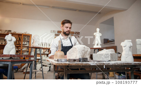 Male sculptor in white shirt and black apron makes a limestone copy of woman torso at the artistic studio. 108193777