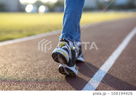 Close up of woman feet in sport sneakers and blue jeans on running lane on outdoor sports court. Close up of woman feet in sport sneakers and blue jeans on running lane on outdoor sports court. 108194926