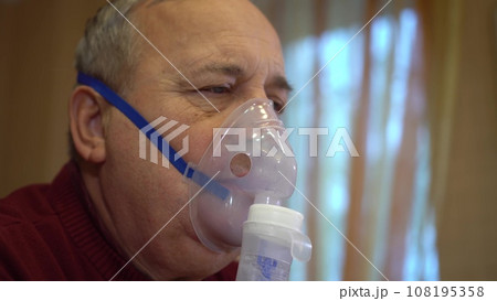 An elderly man inhales through an oxygen mask. A man does a respiratory procedure at home. Closeup. An elderly man inhales through an oxygen mask. A man does a respiratory procedure at home. Closeup. 108195358