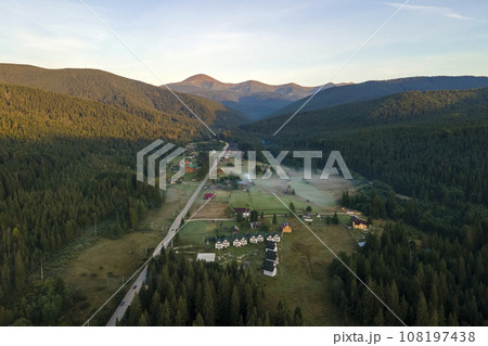 Aerial view of bright foggy morning over small rural homes between dark peaks with mountain forest trees at autumn sunrise. Beautiful scenery of wild woodland at dawn 108197438