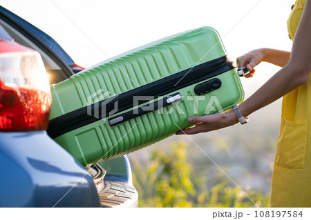 A woman in yellow dress taking green suitcase from car trunk. Travel and vacations concept. 108197584