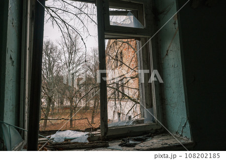 Old broken metal frame window sill of shabby dirty painted walls with black destroyed spider nets and overlooking at green fall trees brick wall building under cloudless blue sky in daytime Old broken metal frame window sill of shabby dirty painted walls with black destroyed spider nets and overlooking at green fall trees brick wall building under cloudless blue sky in daytime 108202185
