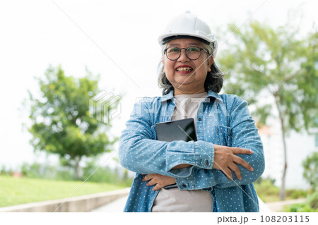 Happy elderly Asian professional engineer wears a helmet and checks the blueprint on a tablet on the construction site after the contractor and architect finish renovation the house and building 108203115