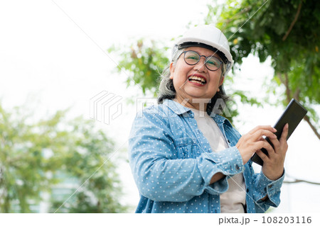 Happy elderly Asian professional engineer wears a helmet and checks the blueprint on a tablet on the construction site after the contractor and architect finish renovation the house and building 108203116
