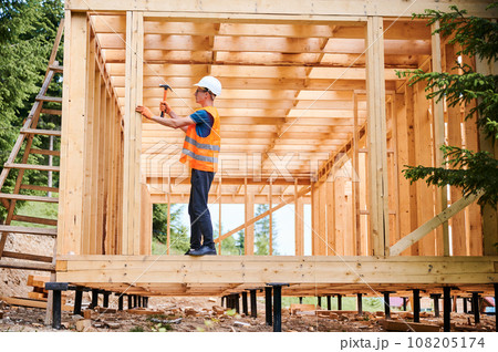 Wooden frame house being built by carpenter. Man wearing glasses hammering nails using hammer and is dressed in a protective helmet and a construction vest. Concept of modern ecological construction. Wooden frame house being built by carpenter. Man wearing glasses hammering nails using hammer and is dressed in a protective helmet and a construction vest. Concept of modern ecological construction. 108205174