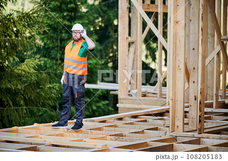 Carpenter constructing wooden frame two-story house near the forest. Bearded man with glasses dressed in protective helmet and orange safety vest. Concept of ecological modern construction. Carpenter constructing wooden frame two-story house near the forest. Bearded man with glasses dressed in protective helmet and orange safety vest. Concept of ecological modern construction. 108205183