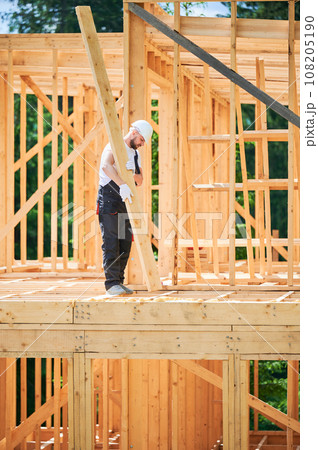 Carpenter constructs wooden-framed house. Man holds large beam in his hands while wearing work clothes and helmet. Idea of modern, eco-friendly construction. Carpenter constructs wooden-framed house. Man holds large beam in his hands while wearing work clothes and helmet. Idea of modern, eco-friendly construction. 108205190