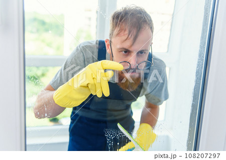 Cheerful bearded man in funny eyeglasses cleans the window with detergents and remove dirt with his finger in gloves. Cheerful bearded man in funny eyeglasses cleans the window with detergents and remove dirt with his finger in gloves. 108207297