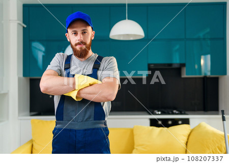 Handsome professional male worker from cleaning service in special uniform and yellow gloves folded his arms on the background of the contemporary cuisine. Handsome professional male worker from cleaning service in special uniform and yellow gloves folded his arms on the background of the contemporary cuisine. 108207337