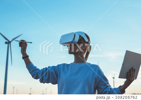 Young woman wearing VR headset while standing on field near windmills Young woman wearing VR headset while standing on field near windmills 108208762