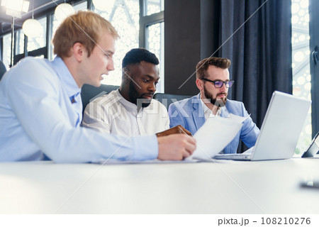 Three mixed races handsome businessmen discussing opportunities of their business strategy using different reports with infographics. Three mixed races handsome businessmen discussing opportunities of their business strategy using different reports with infographics. 108210276