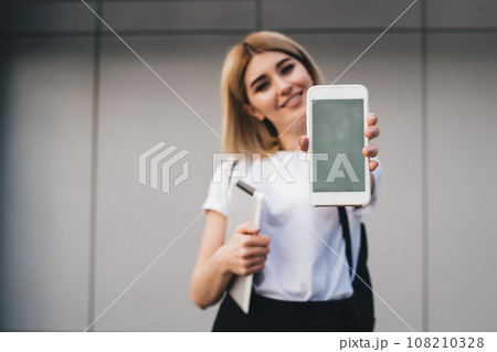 Positive female demonstrating smartphone near office building 108210328