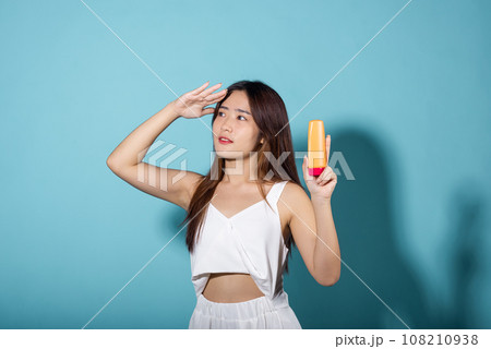 Beautiful Asian young woman holding body lotion on hand in studio shot isolated on blue background, Smiling brunette female hold cream sunblock bottle, tube of sunscreen, protect sun light and UV 108210938