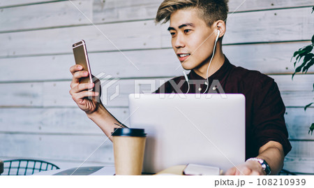 Japanese hipster guy listening music in headphones while checking notification on smartphone device Japanese hipster guy listening music in headphones while checking notification on smartphone device 108210939